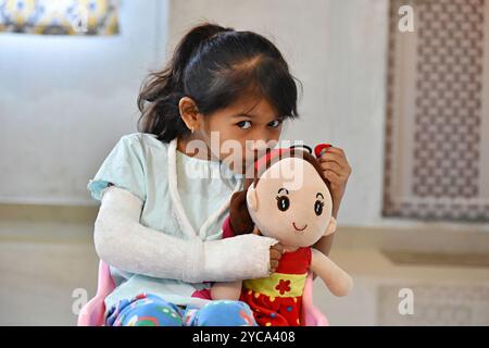 Porträt eines jungen indischen Mädchens mit gebrochener Hand im Gips, das auf einem Stuhl sitzt und ihre Puppe, Pune, Maharashtra, Indien, küsst. Stockfoto