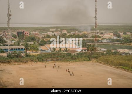 Die afrikanischen Kinder spielen Fußball auf dem staubigen Feld in Banjul, mit einem Luftpanorama der Stadt Gambia. Stockfoto