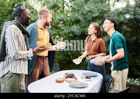 Seitlicher Blick auf eine Gruppe glücklicher Erwachsener, die gemeinsam im Freien einen Tisch legen, um das Essen an grünen Bäumen im Hof zu genießen Stockfoto