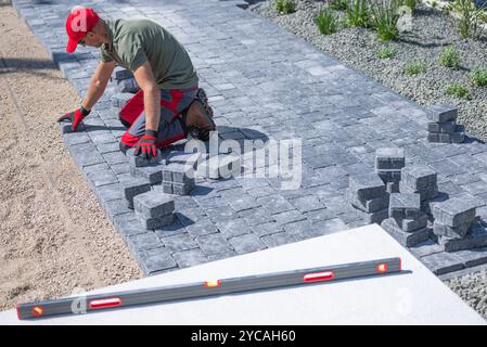 Eine Person kniet auf einem Weg und legt vorsichtig Pflastersteine in einen Wohngarten, umgeben von Grün. Stockfoto