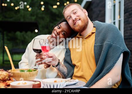 Lebendiges Porträt eines multiethnischen erwachsenen Paares, das sich für ein Foto ausgibt und am Esstisch sitzt und draußen Gläser hält Stockfoto