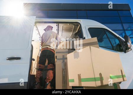Ein Mitarbeiter in lässiger Kleidung entlädt große Kartons aus einem Van, der in einem modernen Büro geparkt ist. Die Sonne scheint hell und wirft Schatten. Stockfoto