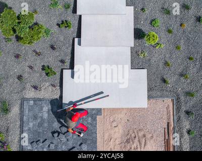 Ein Bauarbeiter platziert vorsichtig Betonstufen in einem Gartenbereich, umgeben von Kies und Pflanzen, tagsüber unter klarem Himmel. Stockfoto