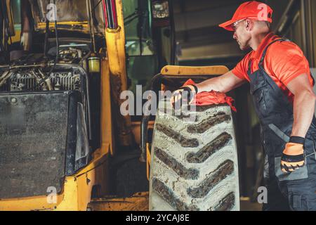 Ein Mechaniker inspiziert einen großen Reifen an einer Baumaschine und konzentriert sich dabei auf Reparaturen, während er eine orangefarbene Kappe und Handschuhe an einer gut beleuchteten Stelle trägt Stockfoto