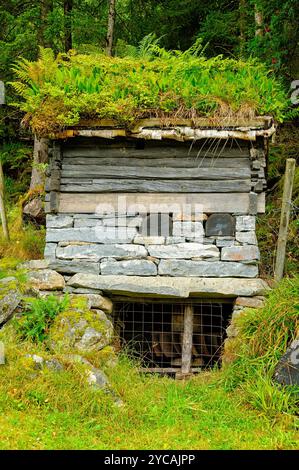 Ein kleines Mühlenhaus mit einem grünen, moosigen Dach und einem Steinfundament befindet sich in einem bewaldeten Bereich. Die Kabine hat ein Metallgitter, das den Eingang abdeckt. Stockfoto