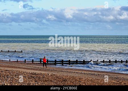 Eine einsame Frau in rotem Mantel, die an einem sonnigen Herbsttag am Strand von Littlehampton entlang läuft Stockfoto