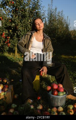 Stilvolles Mädchen in schwarzer Kleidung, das neben gepflückten Äpfeln in einem sonnigen Herbstgarten sitzt. Stockfoto