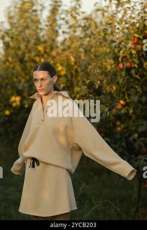 Ein Mädchen in stylischen Trainingsanzügen posiert in einem Apfelgarten bei Sonnenuntergang. Das Model trägt einen hellen Trainingsanzug und Rock. Stockfoto