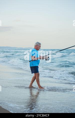 Ein älterer Mann fischt ruhig im Sand von Valencia, Spanien, lässig gekleidet, konzentriert sich intensiv auf seine Angelrute inmitten der sanften Strandwellen Stockfoto