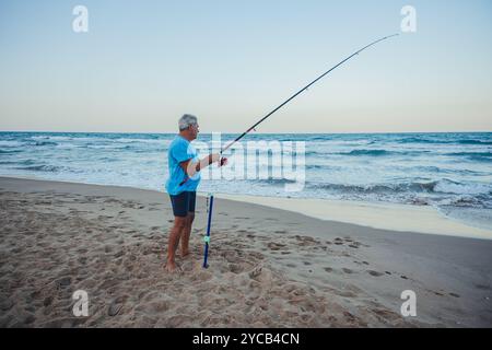 Ein älterer Mann in einem hellblauen Hemd steht beim Angeln am Sandstrand von Valencia, Spanien, während die Wellen in der Abenddämmerung sanft hinter ihm abstürzen Stockfoto