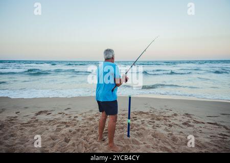 Ein älterer Fischer steht an einem Sandstrand in Valencia, Spanien, und fischt an einem ruhigen Abend im Mittelmeer Stockfoto