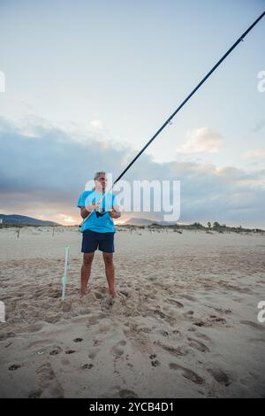 Ein älterer Mann steht bei Sonnenuntergang an einem ruhigen Sandstrand in Valencia, Spanien, fischen. Sein fokussierter Ausdruck und die ruhige Umgebung erwecken ein Gefühl für Stockfoto