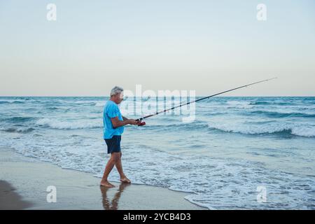 Ein älterer Fischer steht an den Sandstränden von Valencia, Spanien, und handhabt geschickt eine Angelrute, während die Wellen sanft um ihn herum brechen Stockfoto