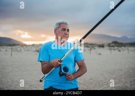 Ein reifer Mann steht an einem Sandstrand in Valencia, Spanien, mit einer Angelrute, mit einem ruhigen Sonnenuntergang und Bergen im Hintergrund Stockfoto