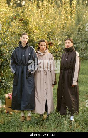 Drei stylische Mädchen in Regenmänteln in einem Apfelgarten. Eine Modenschau im Garten bei Sonnenuntergang. Stockfoto