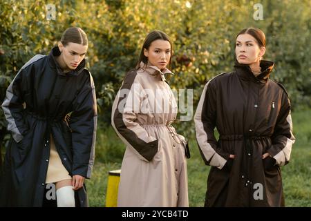 Drei stylische Mädchen in Regenmänteln in einem Apfelgarten. Eine Modenschau im Garten bei Sonnenuntergang. Stockfoto