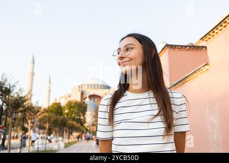 Eine junge Frau, die wegblickt und lächelt, schlendert in die Nähe der berühmten Hagia Sophia im historischen Sultanahmet-Viertel von Istanbul und verkörpert eine entspannte und fröhliche Atmosphäre Stockfoto