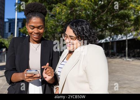 Eine afroamerikanische Frau und eine lateinamerikanische Frau aus lateinamerika, die einen sonnigen Tag im Freien genießt, eine mit einem Smartphone, beide lächeln und auf den Bildschirm schauen Stockfoto