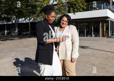 Zwei Geschäftsfrauen, eine Afroamerikanerin und eine hispanistin, die zusammen ein Smartphone benutzen, während sie draußen stehen. Beide lächeln und schauen sich den scr an Stockfoto