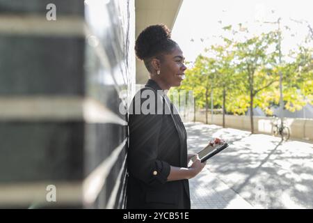 Seitenansicht einer lächelnden afroamerikanischen Geschäftsfrau, die im Freien ein Tablett hält, gekleidet in einem formellen schwarzen Anzug. Sie schaut von der Kamera weg, Stockfoto