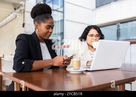 Zwei Berufsfrauen, eine Afroamerikanerin und eine Hispanistin, arbeiten in einem Café. Die Afroamerikanerin schaut auf ihr Smartphone. Stockfoto