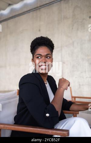 Porträt einer selbstbewussten Afroamerikanerin, die auf einem Stuhl sitzt und wegschaut. Ihr warmes Lächeln und ihre professionelle Kleidung strahlen Selbstvertrauen und Ausgewogenheit aus. Stockfoto