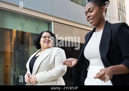 Zwei professionelle Frauen, eine Hispanistin und eine Afroamerikanerin, werden beim Gehen und bei einem angenehmen Gespräch gegen ein städtisches Bürogebäude gesehen Stockfoto