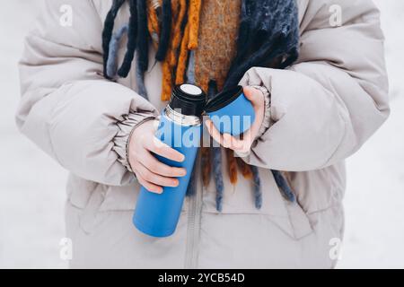 Ein anonymes Teenager-Mädchen in einem warmen Mantel macht einen Winterspaziergang und hält eine leuchtend blaue Thermoskanne. Nahaufnahme von Händen, die ein Getränk ausgießen, ein fördern Stockfoto