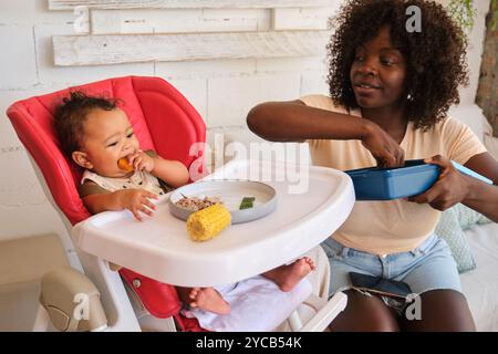 Eine afroamerikanische Mutter füttert ihr Baby gerne in einem roten Hochstuhl, während das Kind ein Stück Mais genießt. Stockfoto