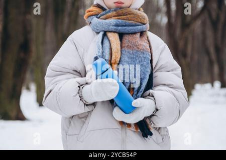 Die abgeschnittene, nicht erkennbare junge Frau in einer Winterjacke und einem Schal hält eine blaue Thermoskanne fest in einem verschneiten Wald. Ihr Gesicht ist nicht sichtbar, was einen Sinn hinzufügt Stockfoto