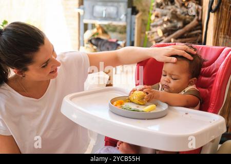 Eine kaukasische Mutter beruhigt sanft ihr afroamerikanisches Baby, das wegschaut, während er in einem Hochstuhl sitzt und unter anderem einen Mais auf dem Kolben erforscht Stockfoto
