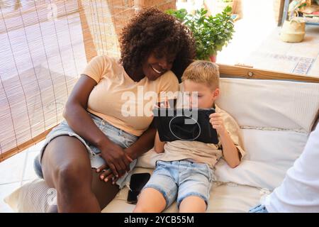 Eine fröhliche Afroamerikanerin und ein konzentrierter kaukasischer Junge sitzen dicht auf einer Couch, vertieft in einer Tablette. Das Licht strömt hinein und sorgt für Wärme im gemütlichen Ambiente Stockfoto