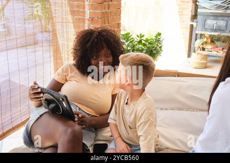 Eine fröhliche afroamerikanische Frau und ein kaukasischer Junge teilen einen zärtlichen Moment, während sie sich auf einem Sofa in einem gemütlichen Raum anschauen, hervorgehoben Stockfoto