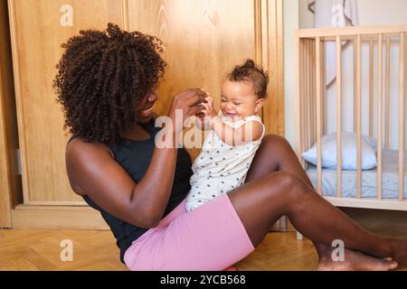 Afroamerikanische Mutter lächelt fröhlich, während sie mit ihrem Baby in einem gemütlichen Zimmer spielt, beide schauen sich neben einem hölzernen Kinderbett an. Stockfoto