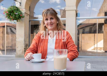 Lächelnde Frau in einer orangefarbenen karierten Jacke sitzt an einem Kaffeetisch und genießt einen Kaffee Stockfoto