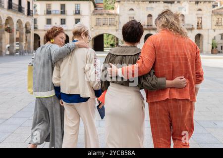 Vier Erwachsene Frauen in stilvollen Outfits umarmen und schlendern auf einem alten Stadtplatz und strahlen ein Gefühl von Freundschaft und Unterstützung aus Stockfoto