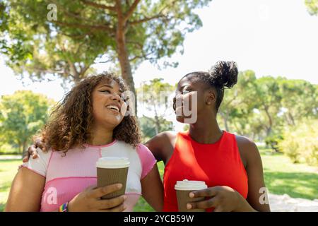 Zwei glückliche Freunde, ein Afroamerikaner und ein Lateinamerikaner, genießen einen sonnigen Tag im Park, teilen sich ein Lächeln und halten Kaffeetassen Stockfoto