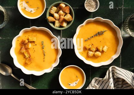 Elegante Kürbissuppe in verschiedenen Schüsseln, abgeschmeckt mit Pfeffer, garniert mit Croutons und frischem Thymian auf dunkler gefliester Oberfläche Stockfoto