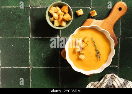 Eine üppige Schüssel cremiger Kürbissuppe garniert mit Croutons und frischem Thymian, serviert auf einem rustikalen Holzschneidebrett über einer grünen gefliesten Oberfläche Stockfoto