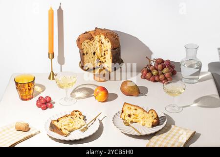 Ein wunderschön präsentiertes italienisches Panettone steht auf einem goldenen Sockel, begleitet von Scheiben auf weißen Tellern, Trauben, Birnen und elegantem Trinkgeschirr und Stockfoto