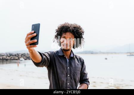 Ein fröhlicher Mann mit lockigen Haaren genießt einen Tag am Strand, hält einen Moment durch ein Selfie mit seinem Smartphone fest und zeigt eine lässige und fröhliche Atmosphäre Stockfoto