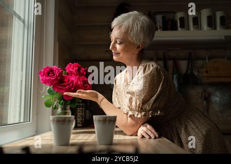 Seitenansicht einer Reifen Frau mit kurzen grauen Haaren lächelt sanft, während sie einen Blumenstrauß von leuchtenden Rosen an einem Fenster in einer rustikalen Küche hält und bewundert Stockfoto