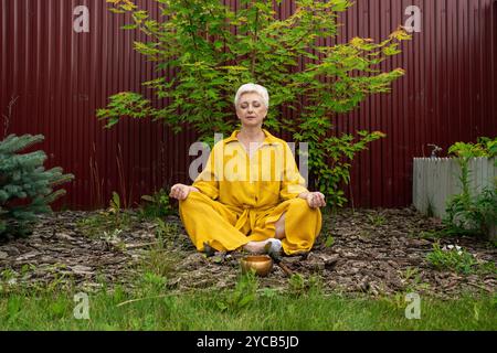 Eine reife Frau in gelbem Outfit meditiert friedlich im Garten mit einer Himalaya-Klangschale. Sie sitzt mit geschlossenen Augen und symbolisiert Stockfoto