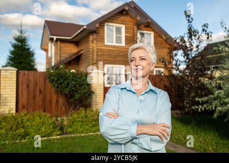 Eine reife Frau mit kurzen, weißen Haaren steht selbstbewusst vor einem rustikalen Holzhaus, umgeben von einem üppig grünen Garten. Sie trägt ein li Stockfoto