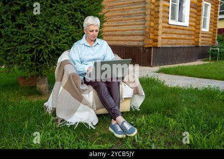 Eine reife Frau sitzt bequem vor einer Blockhütte, mit einer gemütlichen Decke bedeckt, während sie an ihrem Laptop arbeitet, umgeben von üppigem Grün und einem rustikalen Stockfoto