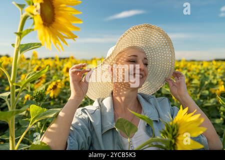 Eine fröhliche reife Frau genießt die Sommersonne auf einem pulsierenden Sonnenblumenfeld, hält sich an ihrem Strohhut fest, mit einem zufriedenen Lächeln und blickt weg vom Stockfoto