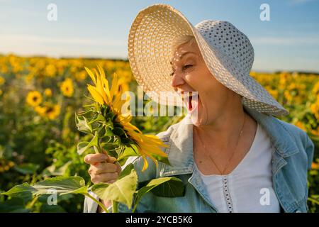 Eine fröhliche reife Frau mit weißem Sonnenhut und Jeansjacke genießt einen sonnigen Tag auf einem weiten Sonnenblumenfeld, hält eine Blüte und lacht glücklich. Stockfoto