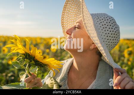 Eine reife Frau, die einen Strohhut trägt, lächelt ruhig, während sie eine Sonnenblume hält. Mit geschlossenen Augen in einem riesigen Feld blühender Sonnenblumen. Das warme Licht Stockfoto