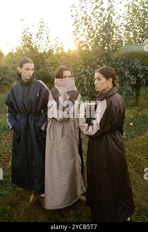 Drei stylische Mädchen in Regenmänteln in einem Apfelgarten. Eine Modenschau im Garten bei Sonnenuntergang. Stockfoto