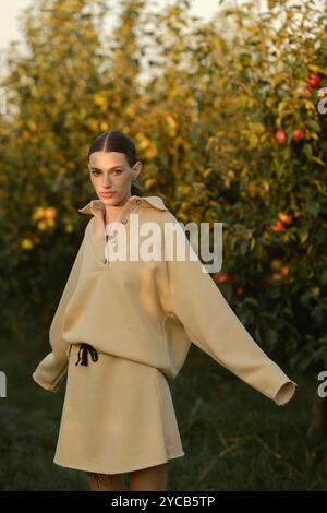 Ein Mädchen in stylischen Trainingsanzügen posiert in einem Apfelgarten bei Sonnenuntergang. Das Model trägt einen hellen Trainingsanzug und Rock. Stockfoto